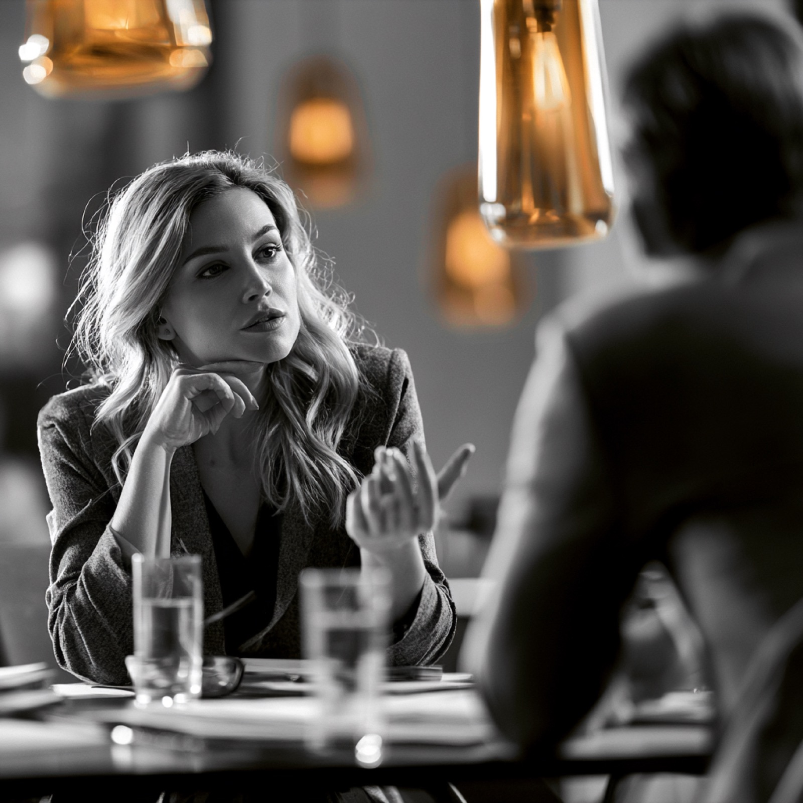 A founder in a grey blazer sits at a table across from a man seen from behind, listening with her chin resting on her hand, glasses of water and papers between them.