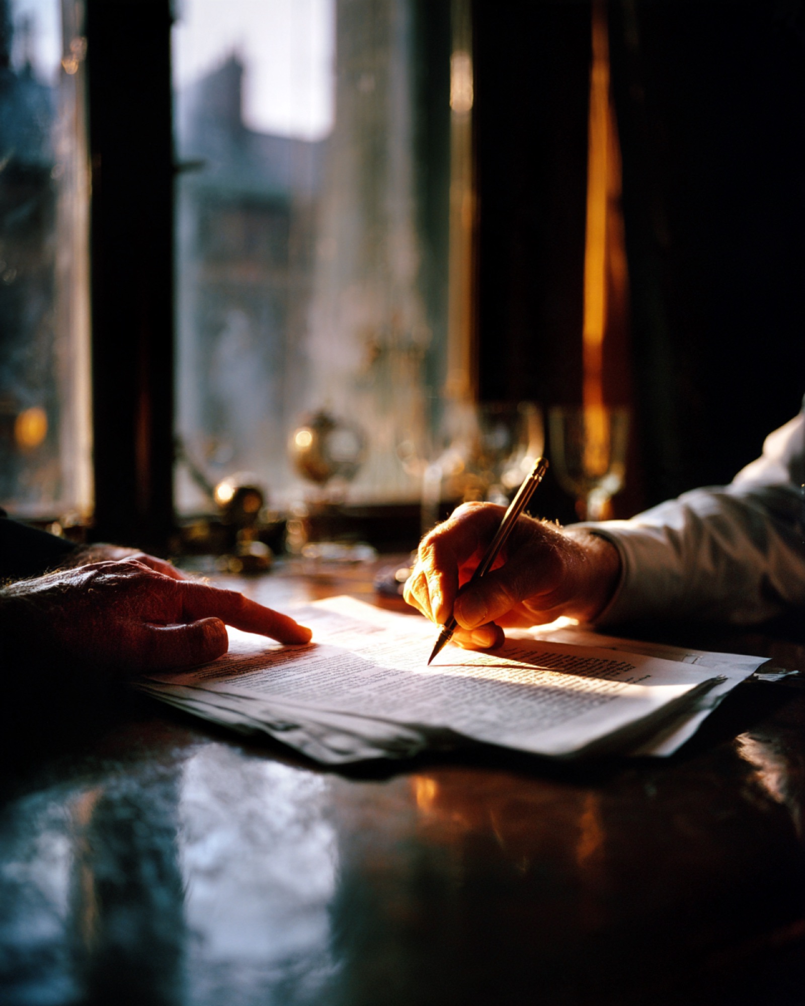 Two people at a polished desk by a window: one points at a line on a printed page while the other marks it with a pen.